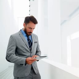 A man in a grey suit and blue shirt stands with a tablet in a bright, modern space.