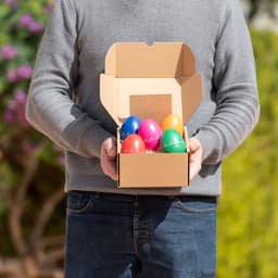A person holding an open cardboard box containing colourful plastic eggs in various bright colours.