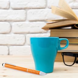 An orange Minox ballpoint pen rests beside a turquoise mug on a wooden surface, with books and glasses in the background.