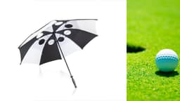 A black and white golf umbrella with circular cutouts, positioned alongside a golf ball on green grass.