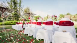 A row of white chairs adorned with red bands, set outdoors amidst flowers and greenery under umbrellas.