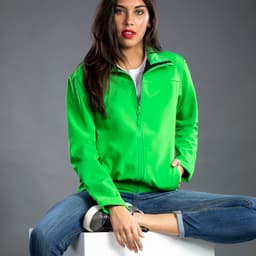 A bright green soft shell jacket with a zip, worn by a model seated on a white cube, featuring a logo.