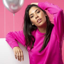 A woman poses in a bright pink hoodie against a pink background, with a silver balloon nearby.