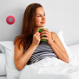 A woman sits in bed holding a green mug, with a pink Bluetooth speaker mounted on the wall.