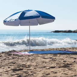 A blue and white beach umbrella standing on sandy shore, with waves gently crashing in the background.