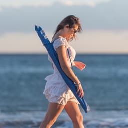 A woman in a white dress walks along the beach, holding a blue Sandok umbrella by her side.