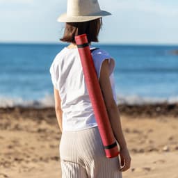 A red and black yoga mat is carried on the back of a woman on a beach, with an ocean backdrop.