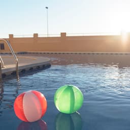Two beach balls, one red and white and the other green, floating in a pool under sunlight.