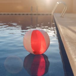 A beach ball in pink and red floats on the water near a poolside, reflecting sunlight.