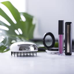A shiny silver hairbrush alongside various beauty products on a marble surface, with green foliage in the background.