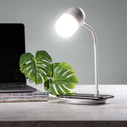 A white multifunction lamp with a flexible neck, wireless charging pad, and a green plant beside a laptop on a desk.