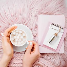 A drink bottle with a metallic finish, placed beside a cup of hot chocolate and a pink notebook on a fluffy surface.