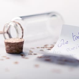 A clear glass wish bottle with a cork stopper, surrounded by scattered glitter and a handwritten note.