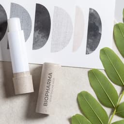 A lip balm in a white and beige tube, accompanied by a decorative card and green leaves. The product has a simple design.