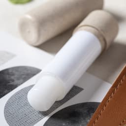 A lip balm with a white stick and a natural-toned cap, placed on a patterned surface with a leather item nearby.