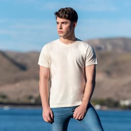 An organic natural adult T-shirt in cream, worn by a model outdoors with a scenic background. The shirt features a logo.