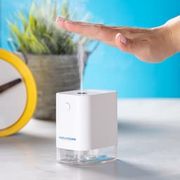 A white hand sanitiser dispenser is emitting a fine mist, set against a blue background and a potted plant nearby.