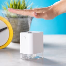 A white hand sanitiser dispenser with a transparent base, positioned on a table next to a potted plant.