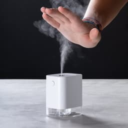 A white hand sanitiser dispenser emits a mist, with a hand positioned above it, set against a dark background.