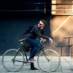 A man on a black bicycle wears a dark sweater and blue jeans, with a backpack and a bag attached to the bike.