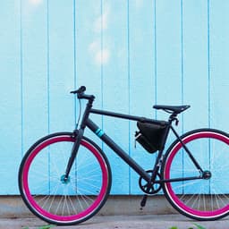 A black bicycle with pink wheels, featuring a small black bike bag attached to the frame against a blue wall.