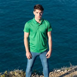 A green polo shirt worn by a model, standing near a coastal area with water in the background. The shirt features a logo.