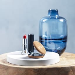 A round pocket mirror in a cork case placed on a white surface, alongside a red lipstick and a blue glass vase.