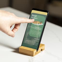 A bamboo phone stand holds a smartphone displaying a recipe page, with a hand touching the screen.
