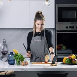 A woman in a grey apron using a knife and wooden spatula in a modern kitchen, with a phone stand beside her.