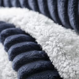 A textured blanket featuring dark navy and fluffy white sections, with a soft, cosy appearance. It has a subtle branding logo.