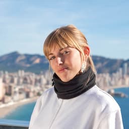 A woman wearing a black scarf stands against a scenic coastal backdrop with mountains and a city skyline.