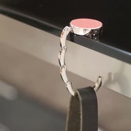 A foldable bag hanger in silver metal with a pink top, designed to clip onto tables or surfaces.
