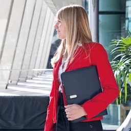 A woman in a red blazer holds a black compendium while standing in a modern office setting with greenery.