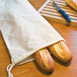 A drawstring bread bag in natural fabric holds two fresh baguettes, with a knife and cutting board in the background.