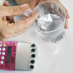 A pink and silver calculator sits next to a clear cup of water, with a hand holding a dropper above it.