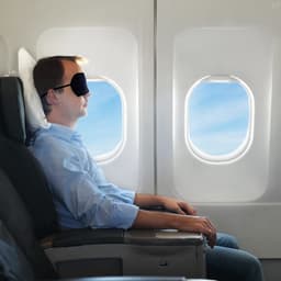 A travel eye mask in black, worn by a man seated by an airplane window, with blue sky visible outside.