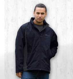 A man wearing a black jacket with a logo, set against a light wood background.