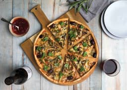 A wooden pizza peel featuring a pizza sliced into pieces, garnished with greens, served with dipping sauce and wine.