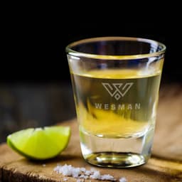 A clear shot glass filled with a yellow liquid, accompanied by a lime wedge and salt on a wooden surface.