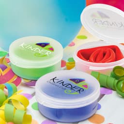 Two containers of colourful, flexible dough in green, red, and blue, with a logo on the lids, surrounded by confetti and balloons.