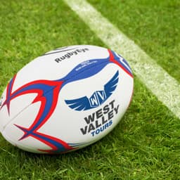 A white touch rugby ball with red and blue accents, featuring a logo on the surface, set on grass.