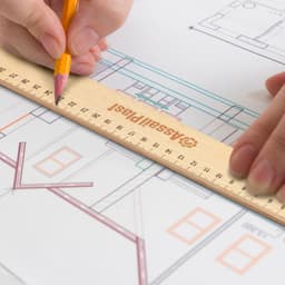 A wooden 30cm ruler rests on a blueprint with a hand holding a pencil. The ruler features a logo.