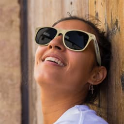 Malibu Basic Sunglasses in natural colour, featuring a logo, worn by a smiling person against a rustic wooden background.