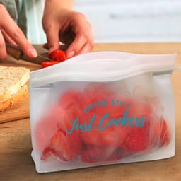 A transparent reusable pouch with strawberries inside, featuring a logo, on a wooden countertop with sliced bread and tomatoes.