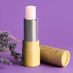 A lip balm in a yellow tube with a silver cap, featuring a light pink balm, alongside lavender flowers.