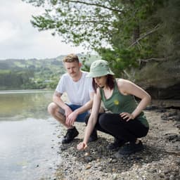 A green tank top is worn by a woman kneeling on the shore, while a man sits beside her. Both are outdoors.