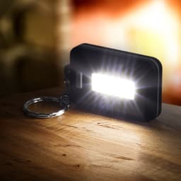 A compact black key ring light emits a bright white light, resting on a wooden surface with a blurred background.