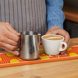 A stainless steel milk jug and a white coffee cup on a vibrant orange PVC bar runner with yellow dots.