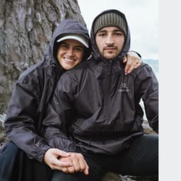 A couple wearing black windbreakers, seated outdoors near a tree, with one jacket featuring a logo.
