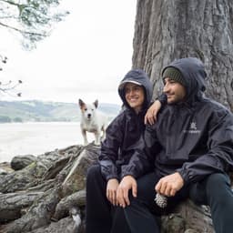 A black windbreaker worn by two people sitting by a tree, with a small dog nearby. The jackets have a logo on them.